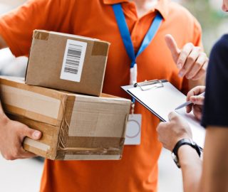 A person wearing an orange T-shirt and a name tag is delivering a parcel to a client, who is putting his signature on the receipt. Friendly worker, high quality delivery service.